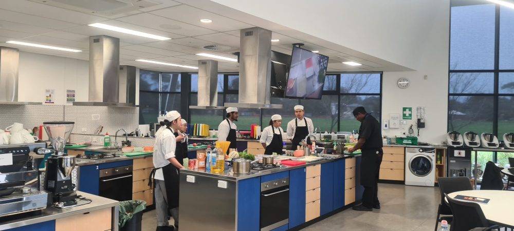 Group of students standing in a kitchen together and learning from the master chef